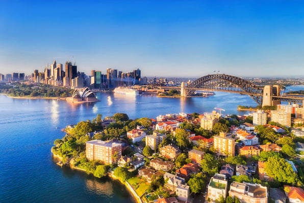 A view of Sydney Harbor including the Sydney Harbor Bridge in the foreground, with the Syndey opera house in the background.